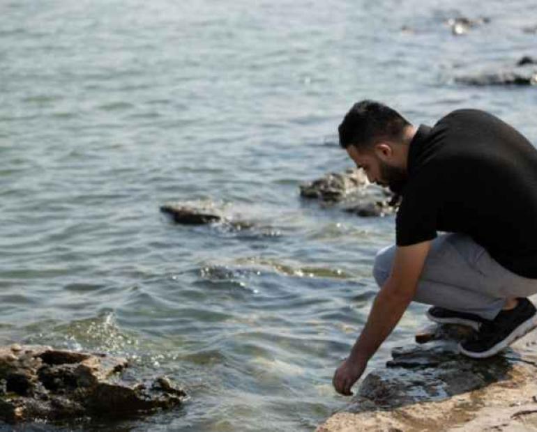 Abdu is crouching by a river, touching the water with his hand.