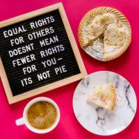 Image is a flat lay from above on a hot pink background. There's a pegboard in the top left showing "Equal rights for others does not mean fewer rights for you - it's not pie" in all caps. The pegboard has a wooden frame and black background with white pegs. In the top right is pie with a slice missing and sprinkles on top. In the bottom right is the slice of pie on a marbled plate. In the bottom left is a white mug filled with coffee.