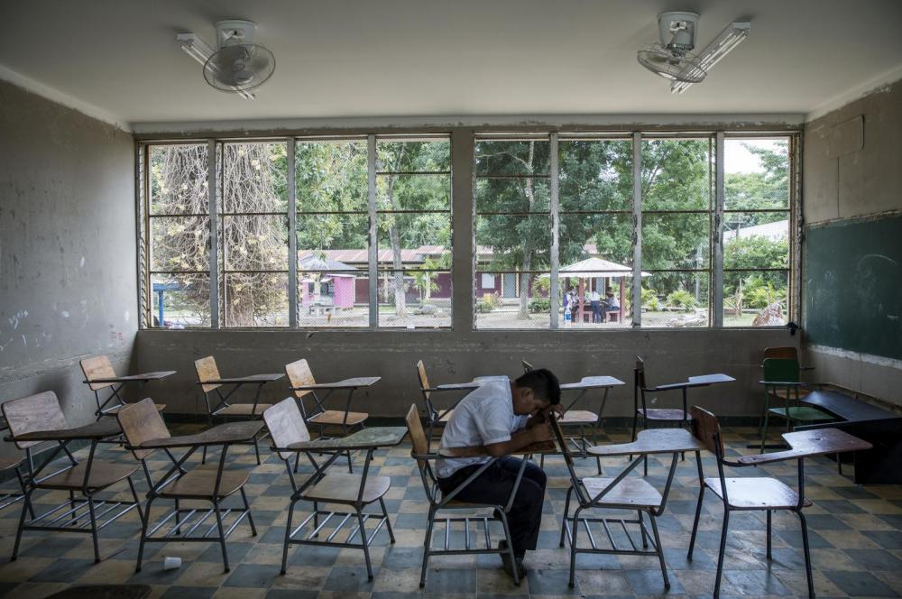 A child sits in an empty classroom with his head on his desk.